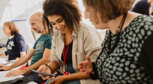 Two people using computers in the background, with two women laughing and looking at a phone in the foreground