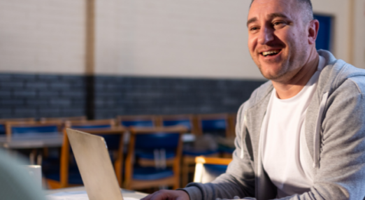 A man smiling and talking to someone while using a laptop in a community centre